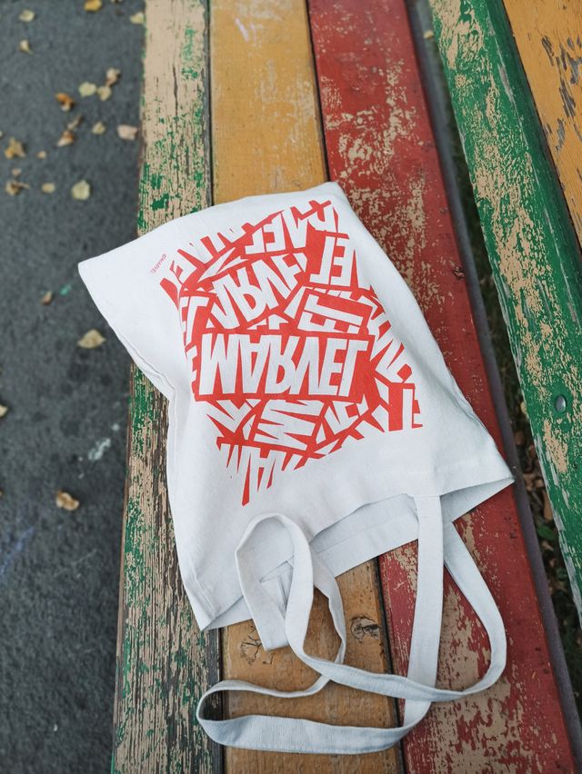 White tote bag with red text placed on a colorful, weathered wooden bench outdoors.