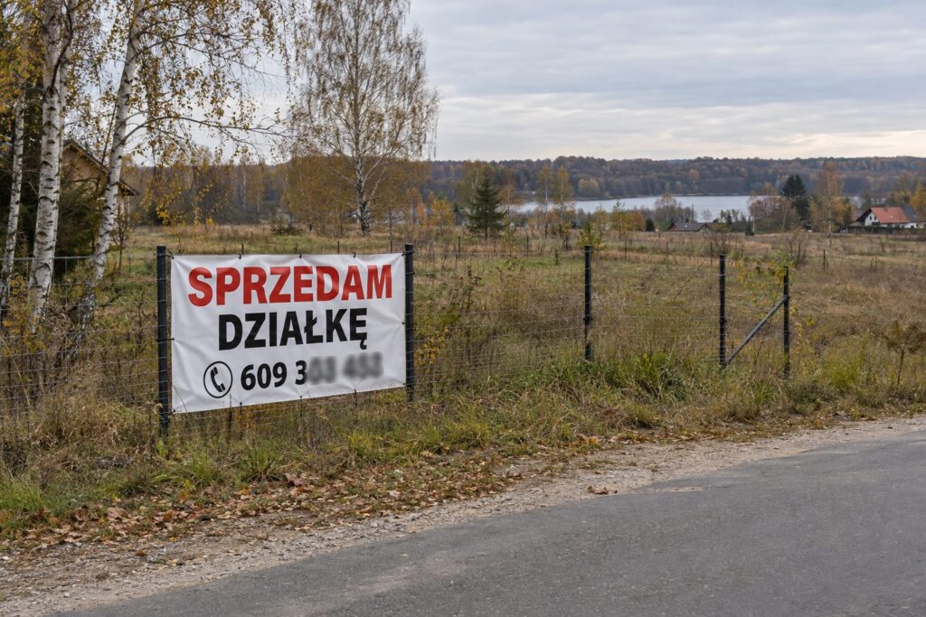 Baner SPRZEDAM DZIAŁKĘ z numerem telefonu na ogrodzeniu działki nad jeziorem