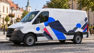 Biały samochód dostawczy z dużymi naklejkami graficznymi na boku, zaparkowany na rynku małego miasta